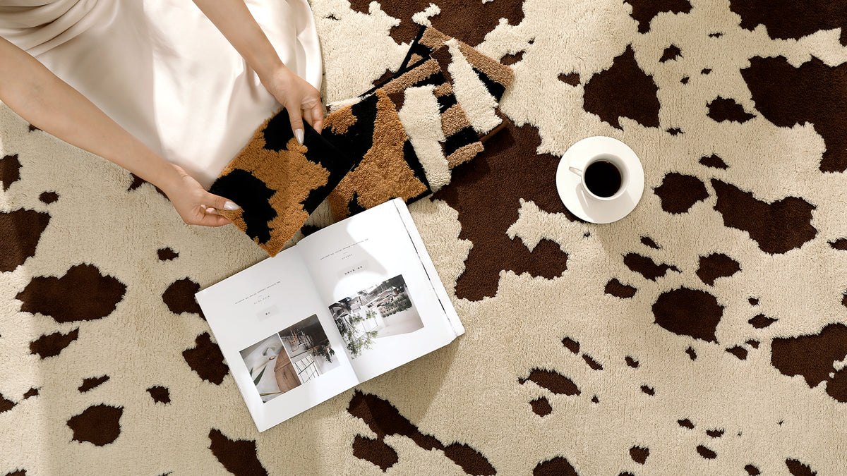 A top-down lifestyle shot of a person sitting on a brown and beige cow-print rug, holding a matching cow-print throw blanket. An open magazine and a cup of coffee are placed nearby on the rug.