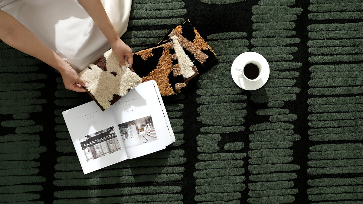 A top-down lifestyle shot of a person holding two textured rug samples on a dark green patterned rug, with an open magazine and a white coffee cup nearby.