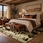 A styled bedroom scene featuring a large cowhide-pattern area rug in beige and dark brown, with a brown leather tufted bench at the foot of the bed. The room has warm wood ceiling beams, a window looking out onto a desert landscape, and a bed dressed in cream bedding with brown and beige accent pillows.