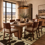 A rustic wooden dining table with six matching wooden chairs featuring cream upholstered seats, arranged on a YIHOUSE cow-print area rug in a dining room. The room has warm beige walls, a large window looking out onto a desert landscape, and a bronze pendant light hanging above.