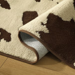 A close-up of a plush area rug with a brown and cream cow-print pattern, shown partially rolled over to reveal the underside and a brown edge, placed on a light wood floor.