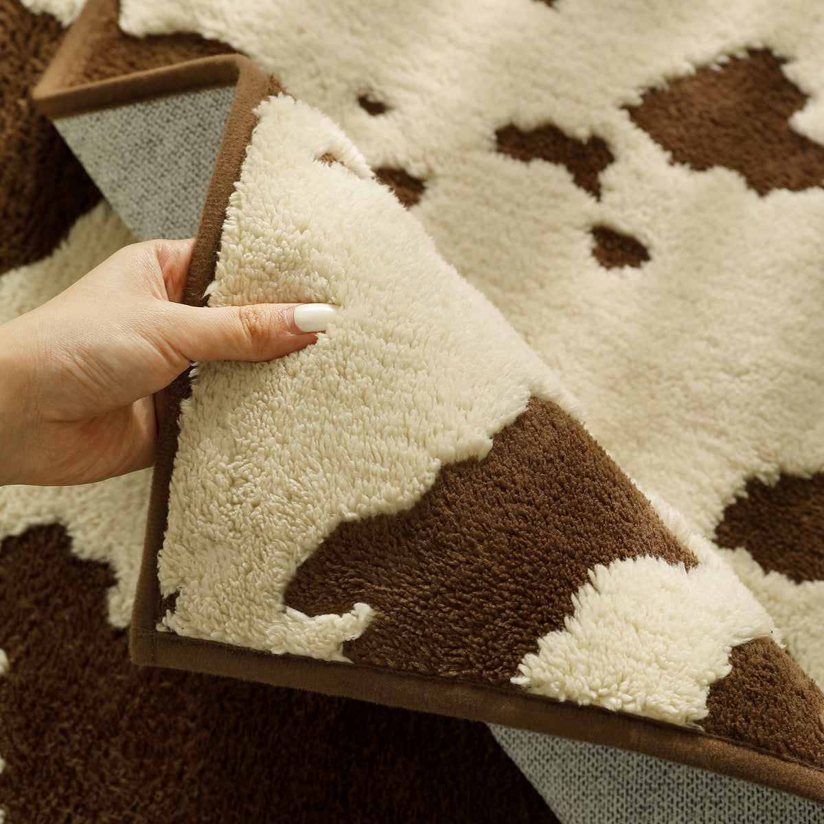 A close-up of a cow-print shag rug being held by a hand to show its thick pile texture. The rug features a cream and brown cow pattern with a solid brown border.