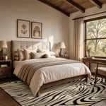A styled bedroom interior featuring a bed dressed in beige and cream linens, with a black-and-cream zebra-pattern rug, wooden ceiling beams, and a large window with sheer curtains.