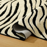 A close-up of a plush zebra-print rug with black and cream stripes, folded over to show its textured backing, placed on a light wood floor.