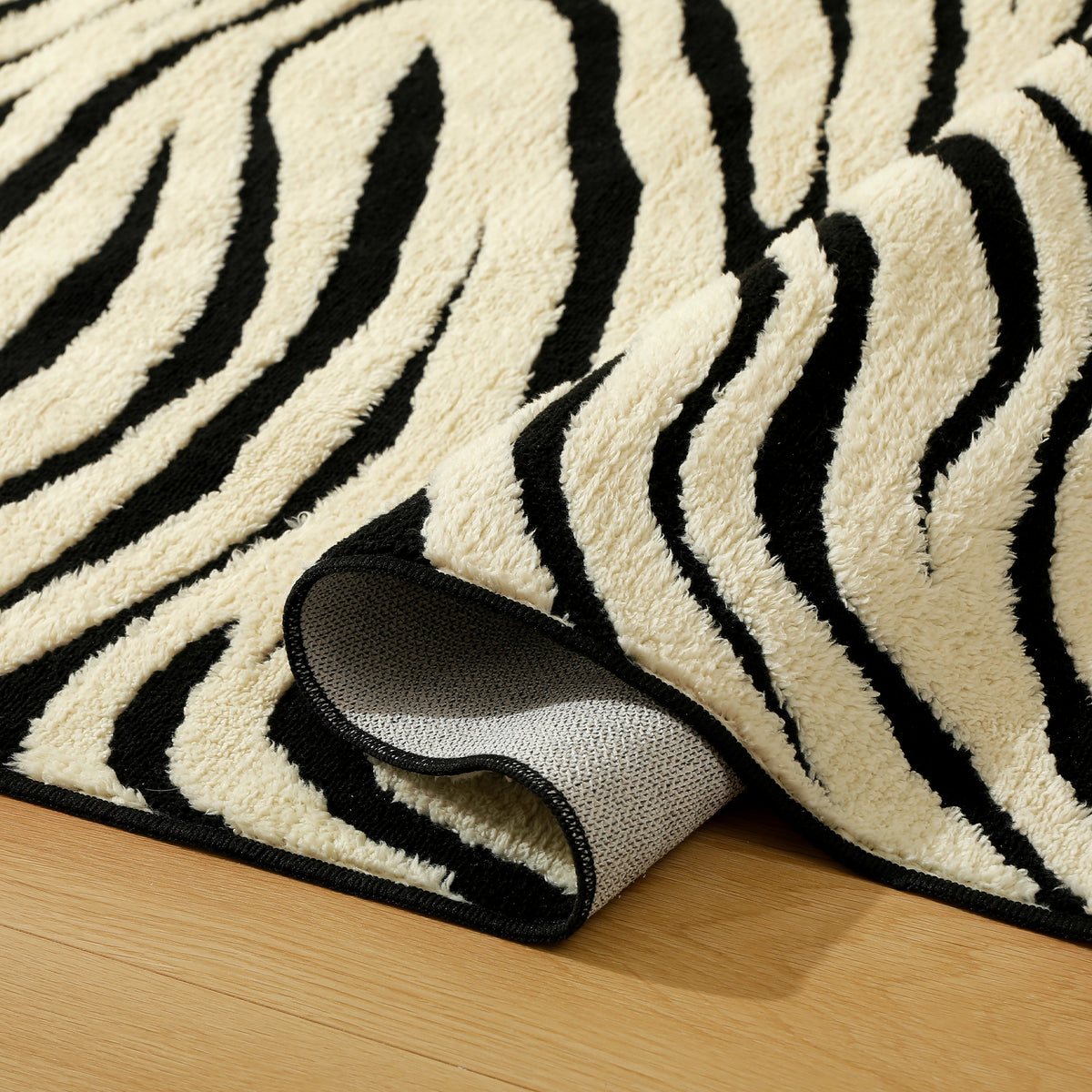 A close-up of a plush zebra-print rug with black and cream stripes, folded over to show its textured backing, placed on a light wood floor.
