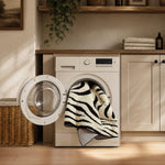 A cream-colored front-loading washing machine stands open in a bright, sunlit laundry room, with a black-and-white zebra-patterned towel draped from the door. Wooden cabinets, a wicker basket, and a hanging plant are visible in the background.
